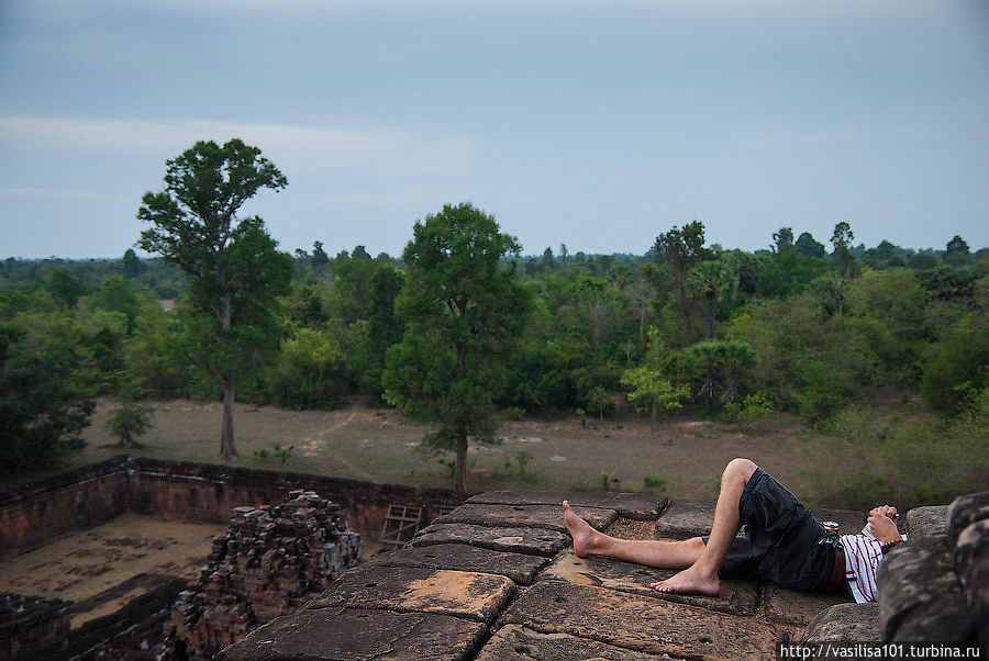 В храме нельзя курить и пить алкоголь. Но хитрый турист спрятался от смотрителей за башней и делает то и другое в ожидании заката. Ангкор (столица государства кхмеров), Камбоджа