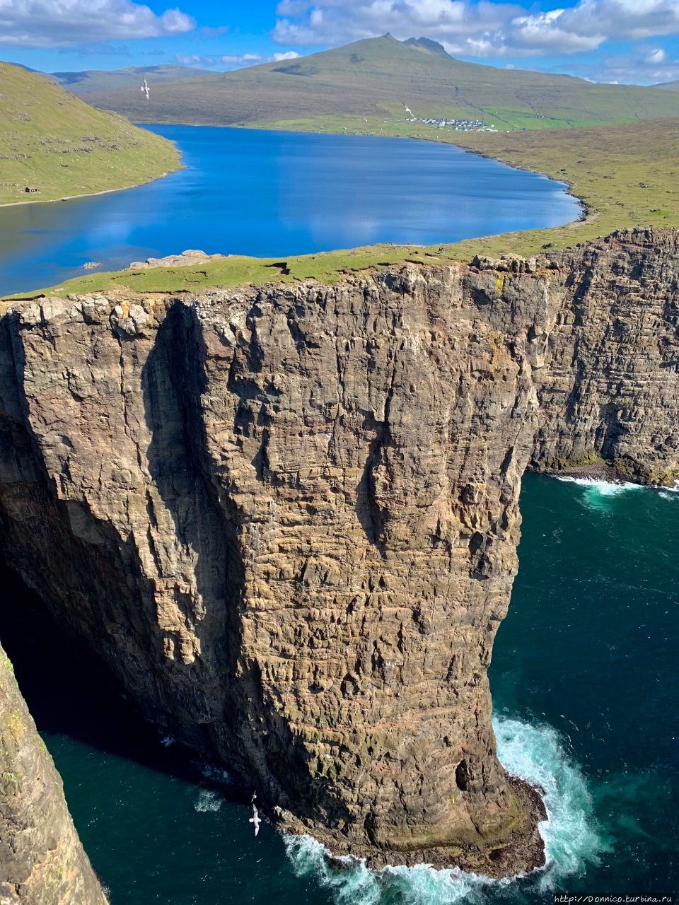 Только c этой точки создается знаменитая оптическая иллюзия Фарерских островов, где кажется, что озеро находится значительно выше своей фактической высоты в 40 метров над уровнем океана...  

Но что еще больше поражает и приводит всех в полный восторг — это цвета озера, изумрудной травы, неба и облаков, и бушующего внизу моря в яркий солнечный день!