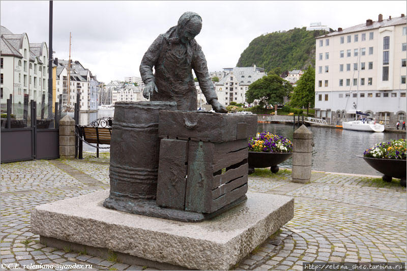 34. А рядом стоит другой памятник, который тоже напоминает о делах прежних веков. Это Sildekona -женщина сельди,  или сельдяная женщина. Скульптура изображает торговку рыбой, а может быть, жену рыбака, которая солит сельдь и укладывает ее в бочку. Эта скульптура отображает реалистичную картину прошлого, когда главной промысловой рыбой и главным товаром тут была сельдь. В те времена такие женщины были типичны для этого городка. Скульптура установлена в 1990 году, деньги на нее дали компании и частные лица. Олесунн, Норвегия