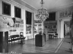 А вот так выглядел этот зал в 1947 году. Это уже экспозиция музея архитектуры. Зал наполнен фотографиями, картинами, фрагментами архитектурных деталей, старинной мебелью. Но роспись потолка уже утрачена, и паркет другой — елочкой.